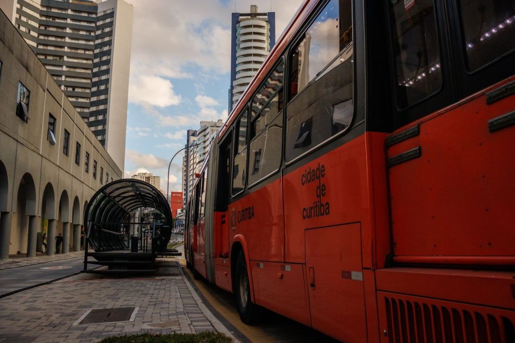 PEC apresentada pelo governo mira transporte gratuito aos fins de semana Ônibus urbano vermelho parado em um ponto moderno no centro de Curitiba.